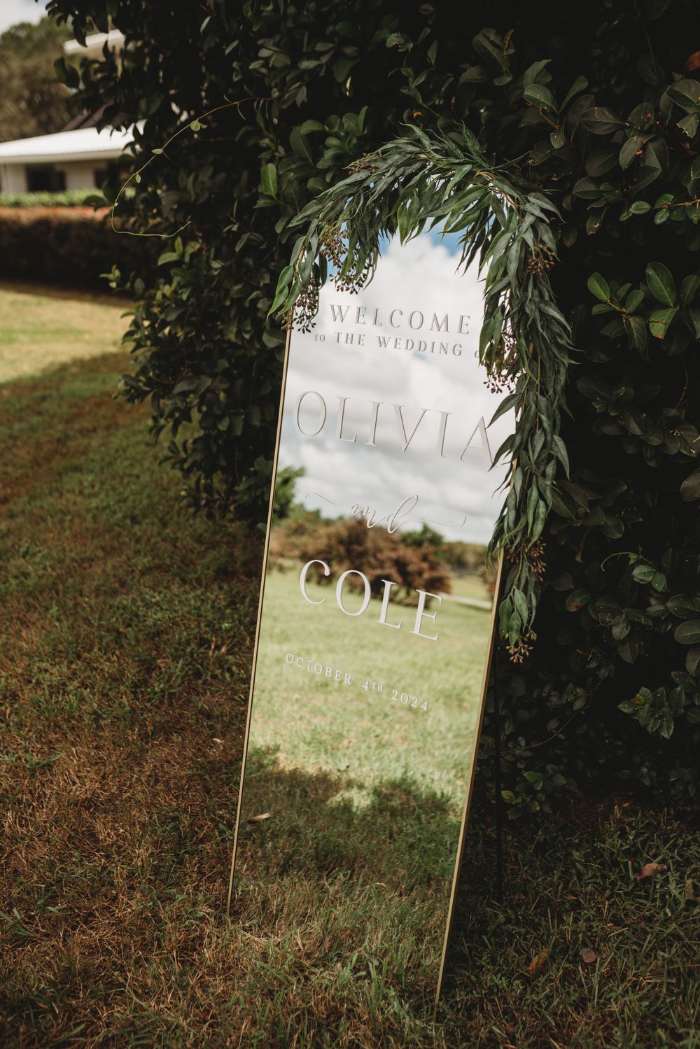 A mirror welcome sign decorated with greenery stands in a garden setting.