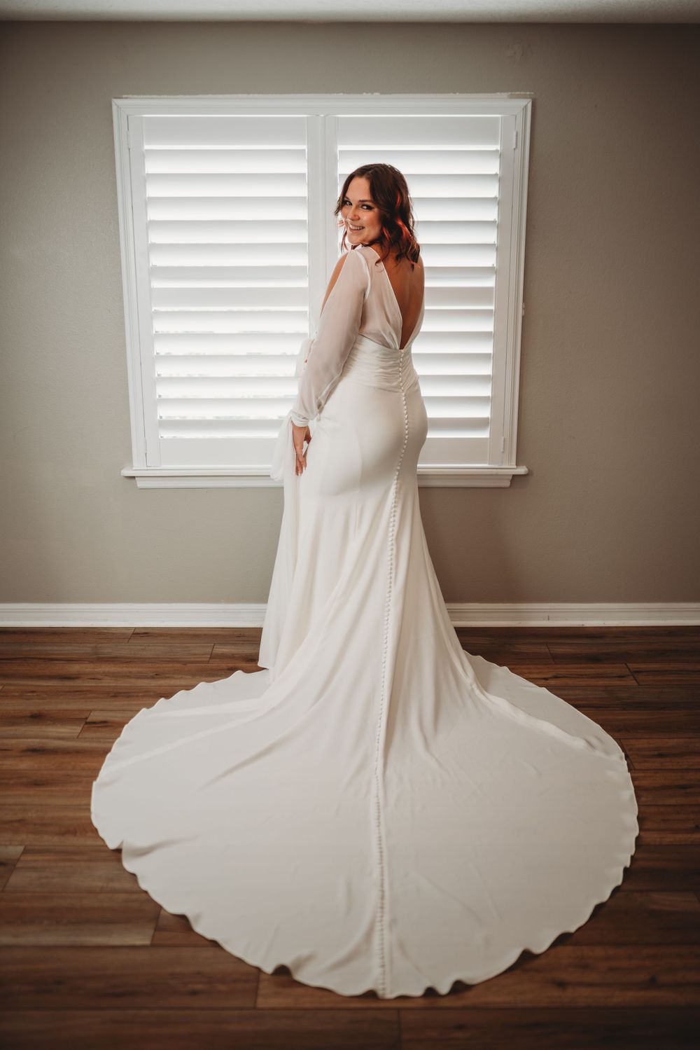 A wedding dress with long sleeves and flowing train photographed against white shuttered window.