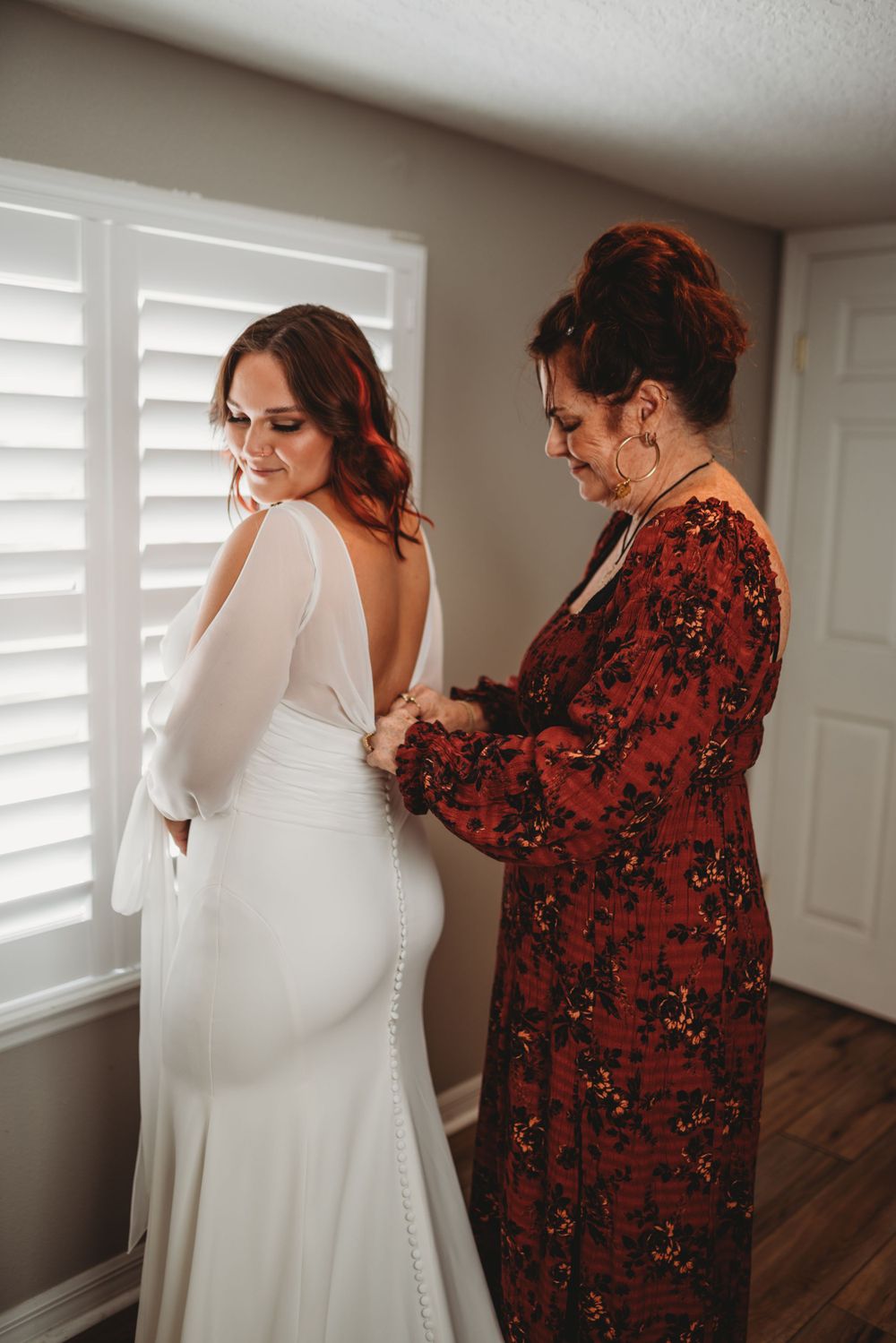 A wedding dress being fastened in a room with white shutters and wooden floors.