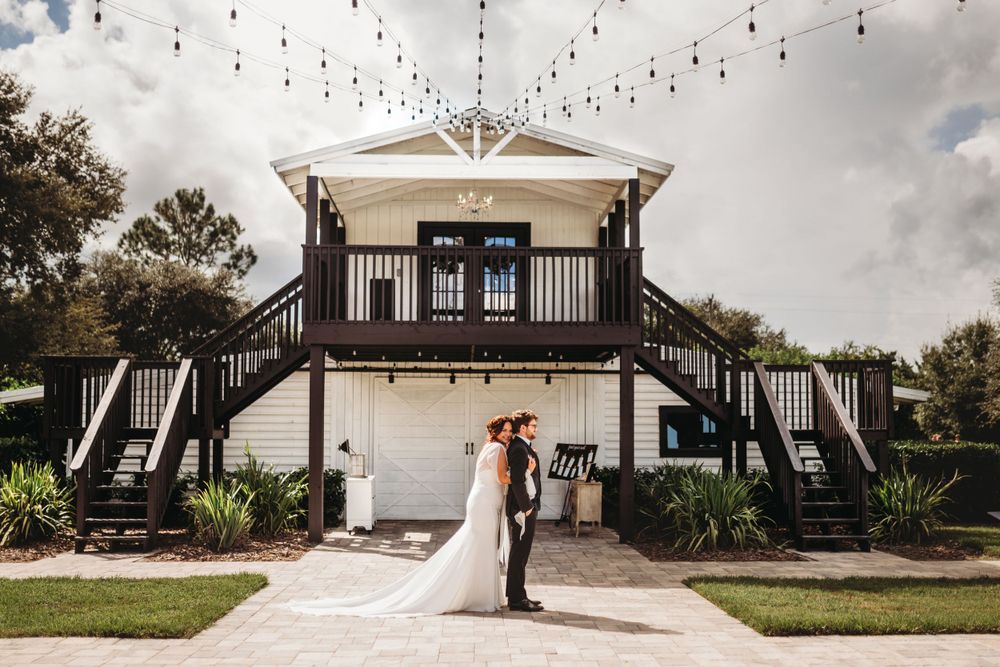A wedding couple poses in front of a white building with symmetrical staircases and string lights overhead.