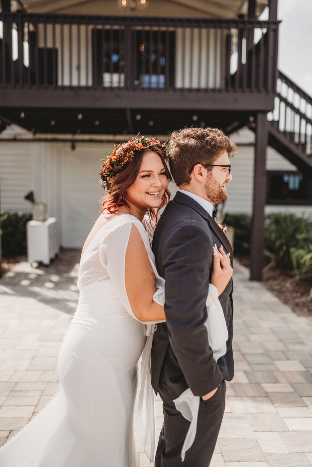 A couple in wedding attire poses back-to-back in front of a white building with dark trim.