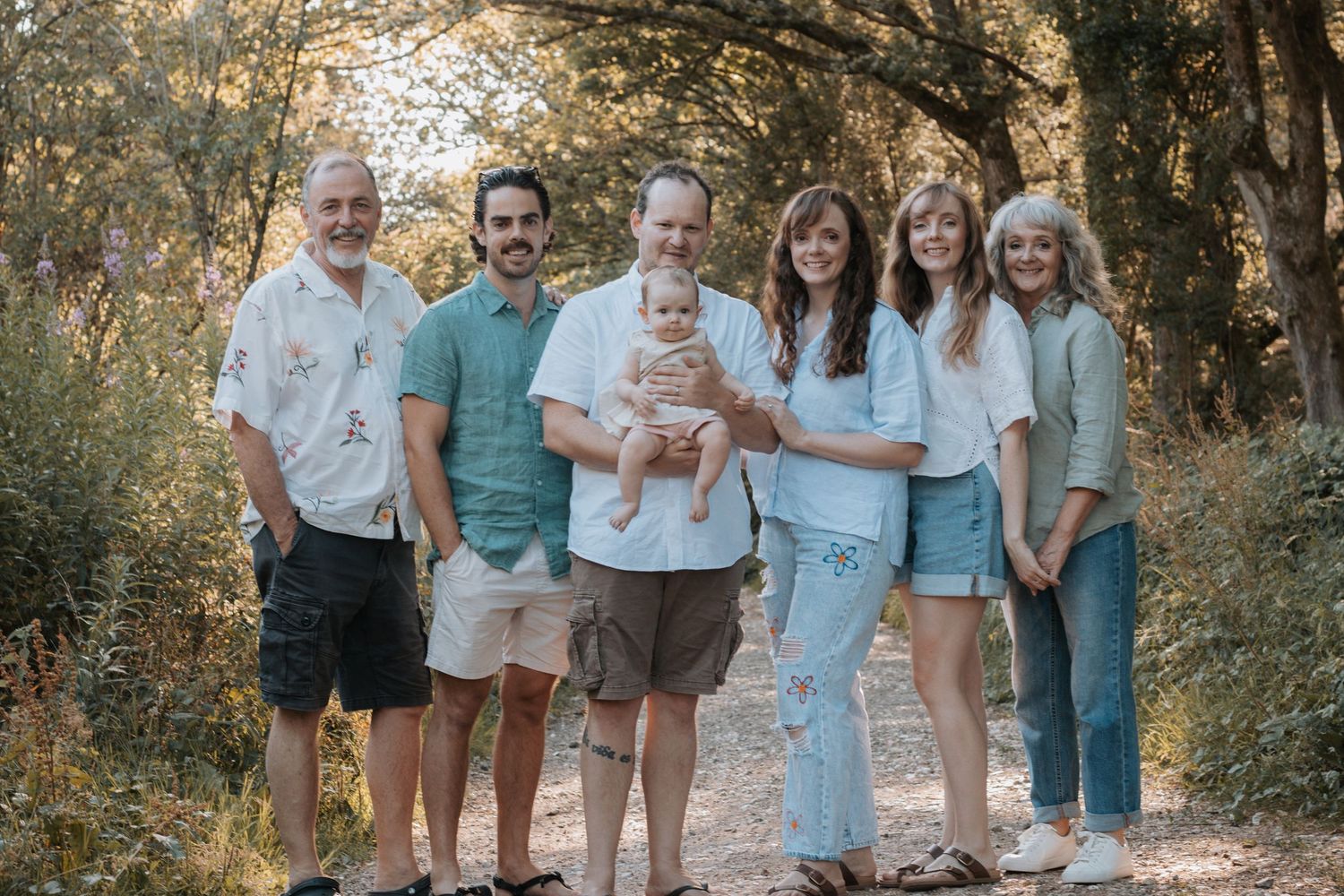 Family of six adults and a baby posing together on a sunny dirt path surrounded by lush green trees.