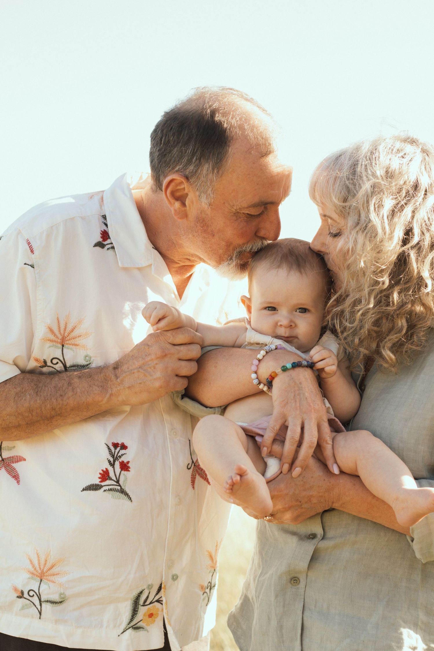 Grandparents lovingly kissing and holding their baby grandchild outdoors in warm sunlight.
