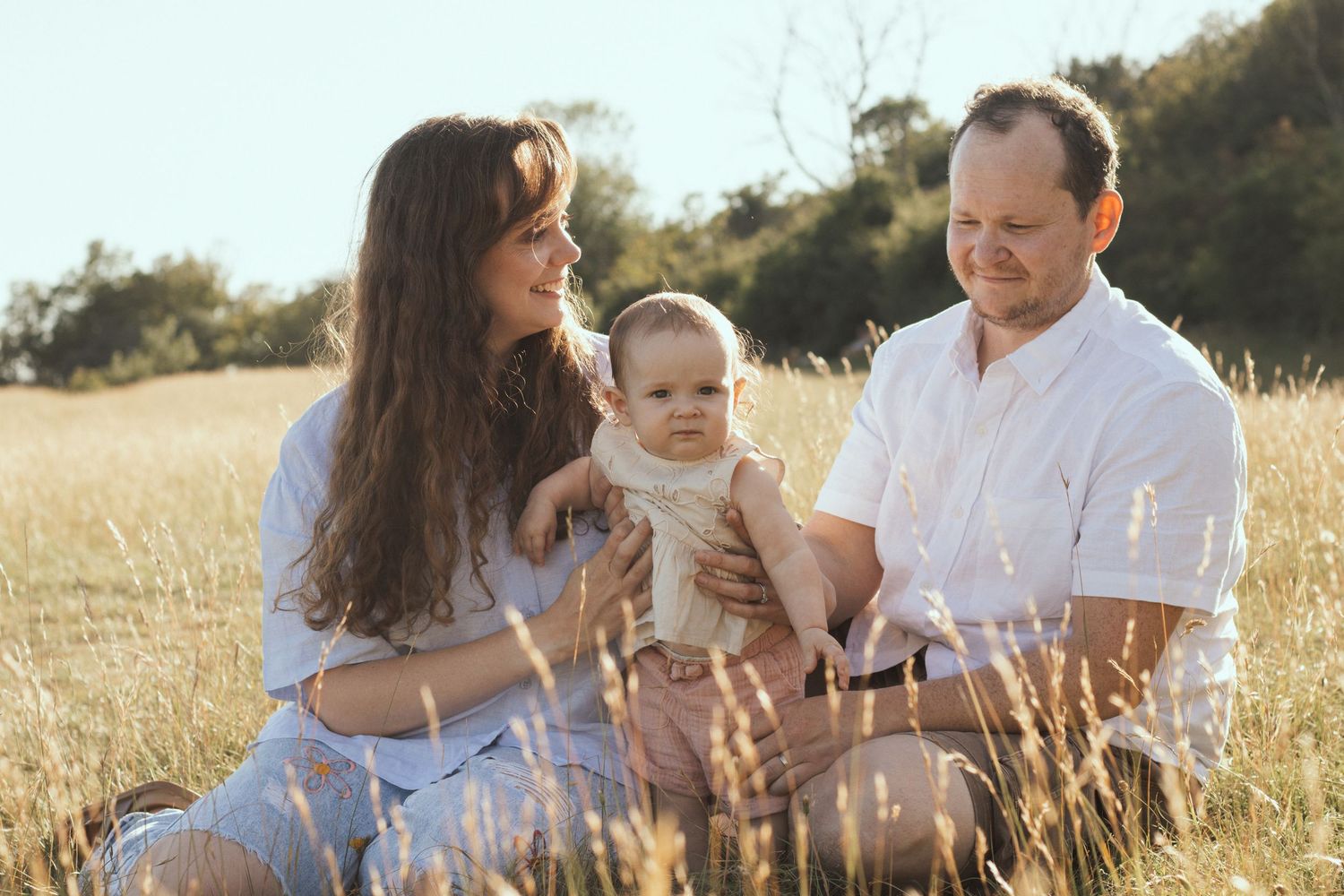 A young family with a baby sits in a golden field, enjoying a warm sunny day outdoors together.