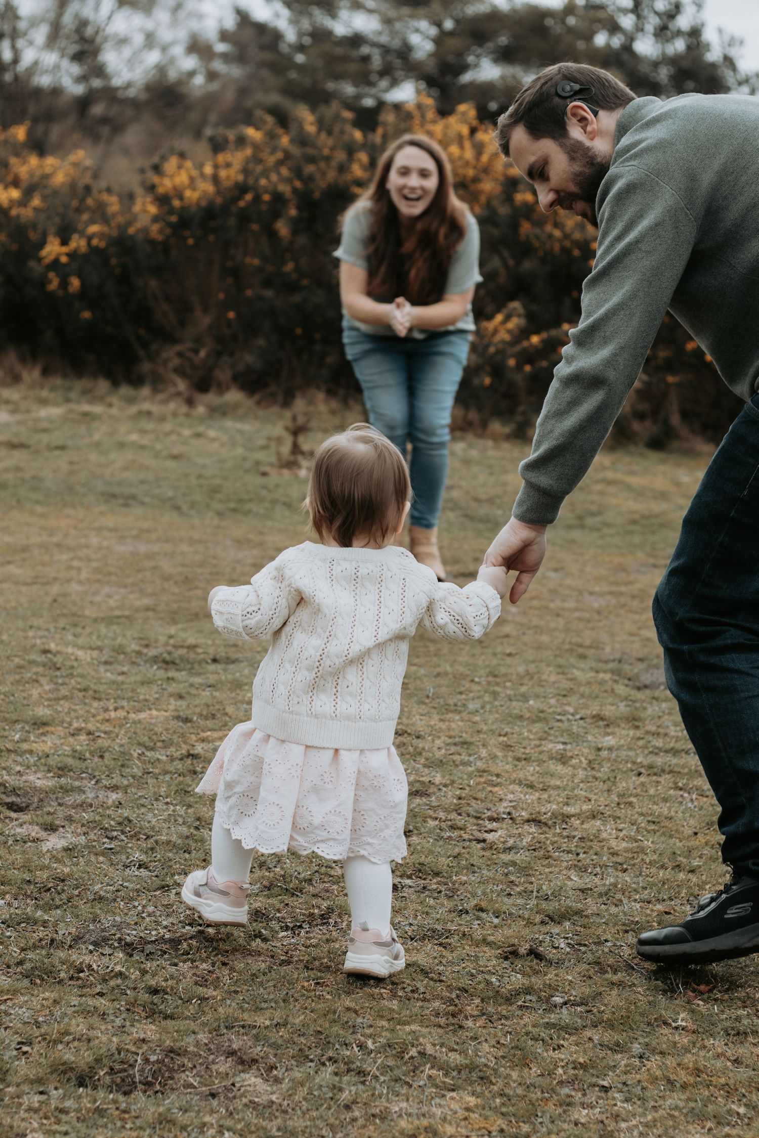 A toddler girl in a white sweater and pink skirt takes first steps toward her laughing mother, guided by her father outdoors.