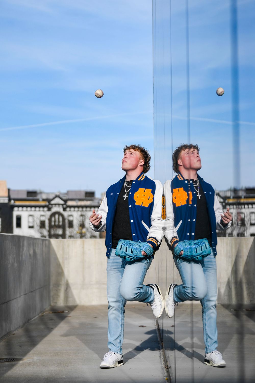 Young man in blue varsity jacket and jeans tosses a baseball beside his mirror reflection on a rooftop.