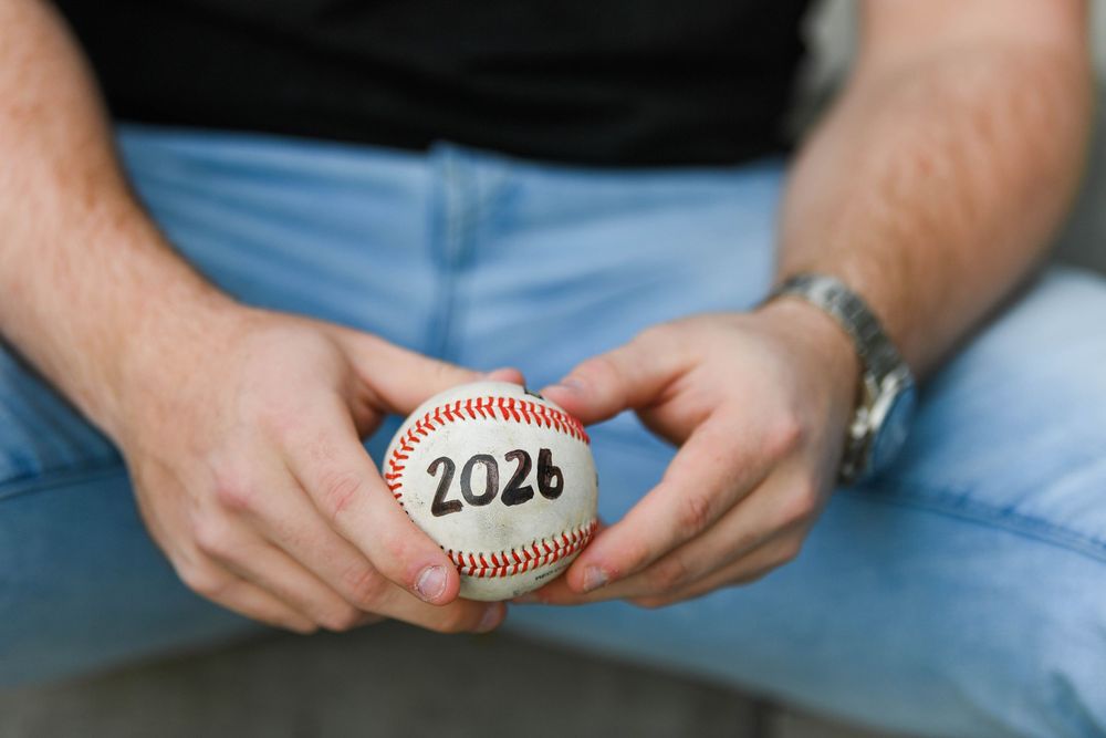 Man holding a baseball with 2026 written on it, wearing jeans and a watch, close-up graduation announcement photo.