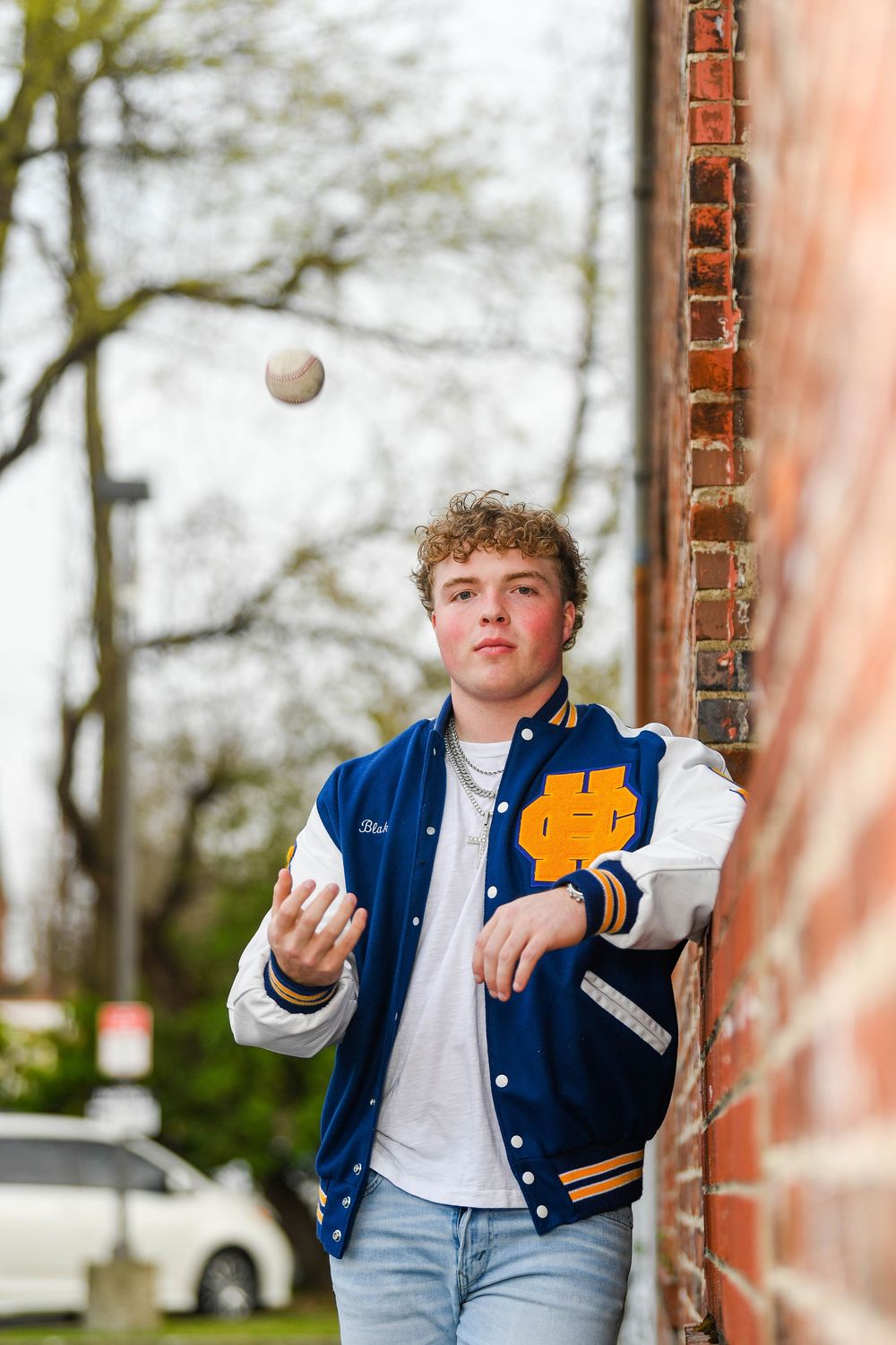 Teen boy in blue varsity jacket tossing a baseball beside a brick wall outdoors.