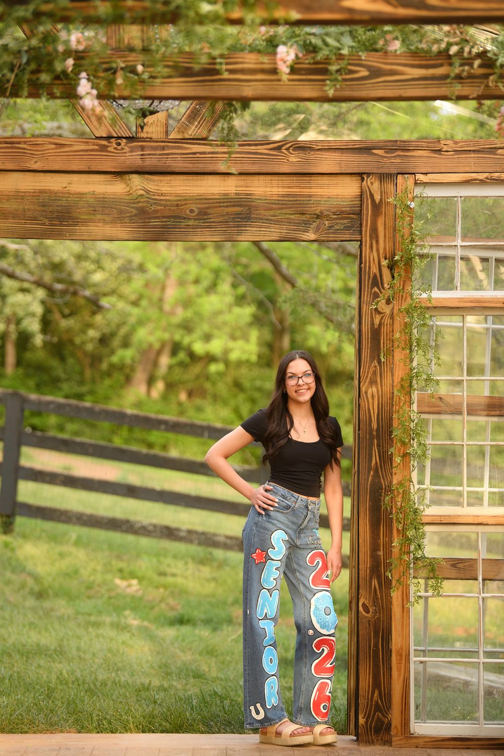 Teen girl in painted Senior 2026 jeans poses under a wooden pergola with greenery at a farm setting.
