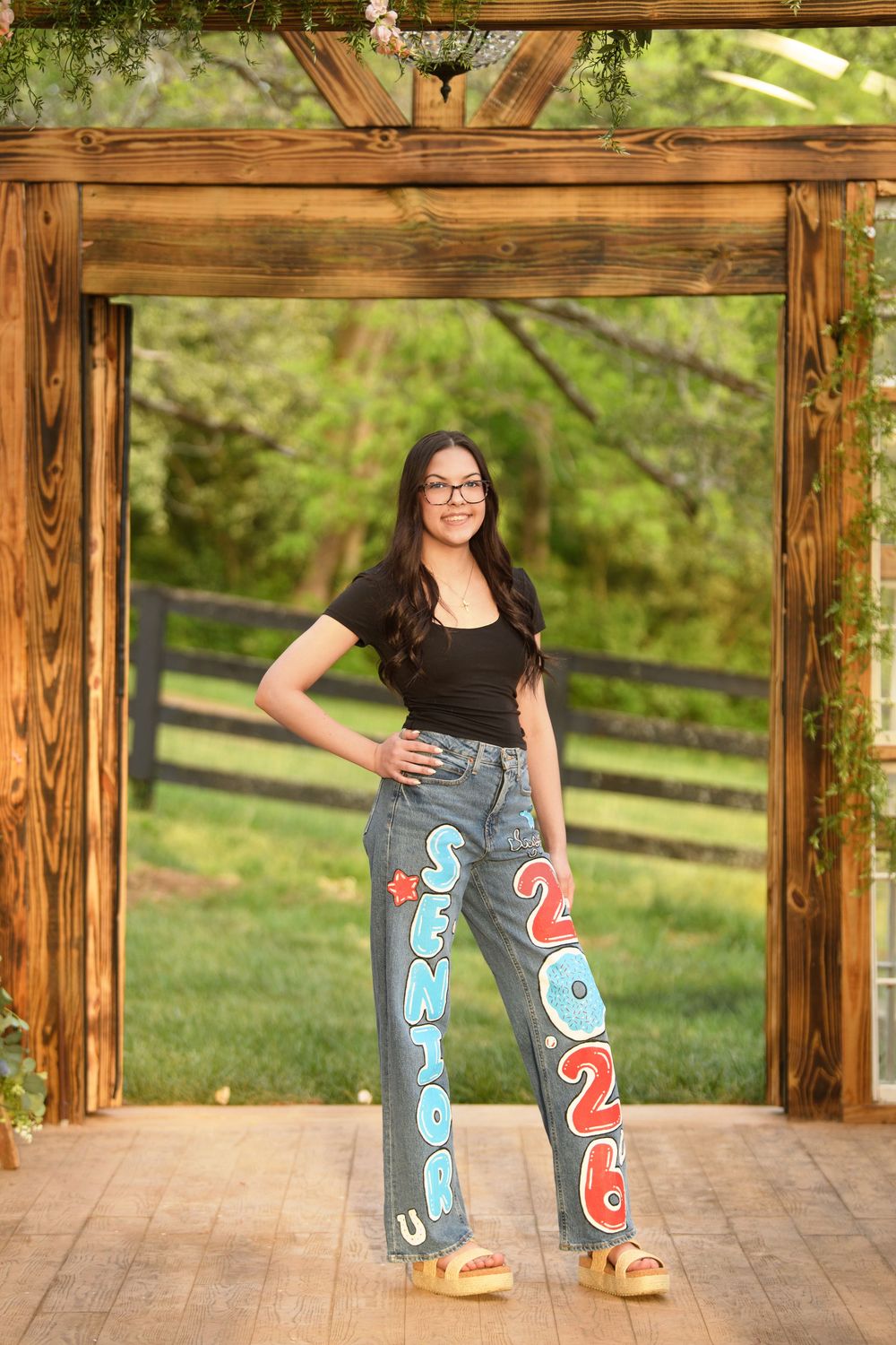 Teen girl poses in custom painted Senior 2026 jeans under a wooden pergola in an outdoor senior portrait.