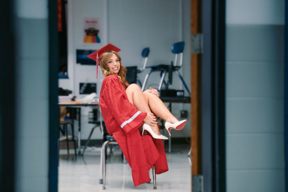 Young woman in red graduation cap and gown sitting in a classroom chair, celebrating her academic achievement.