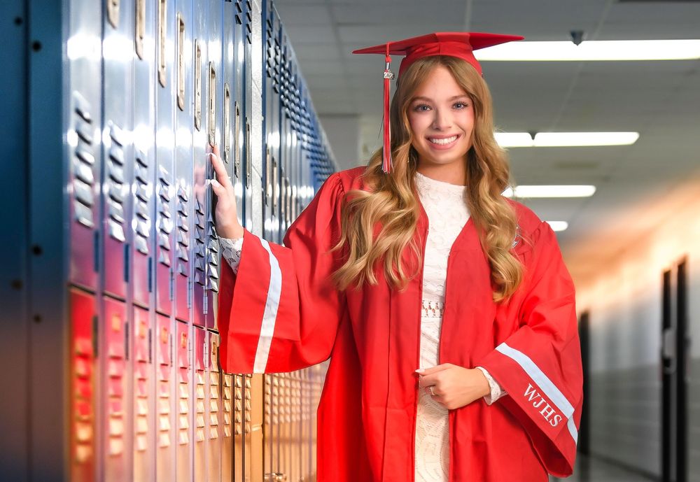Smiling graduate in red cap and gown poses by school lockers, celebrating high school graduation.