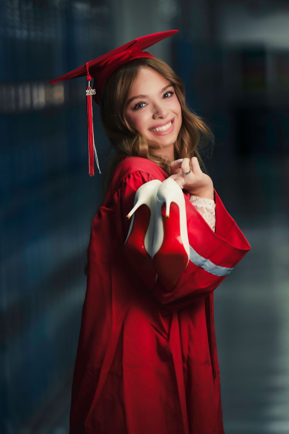 Smiling young woman in red graduation cap and gown holding white heels, posing proudly in a school hallway.