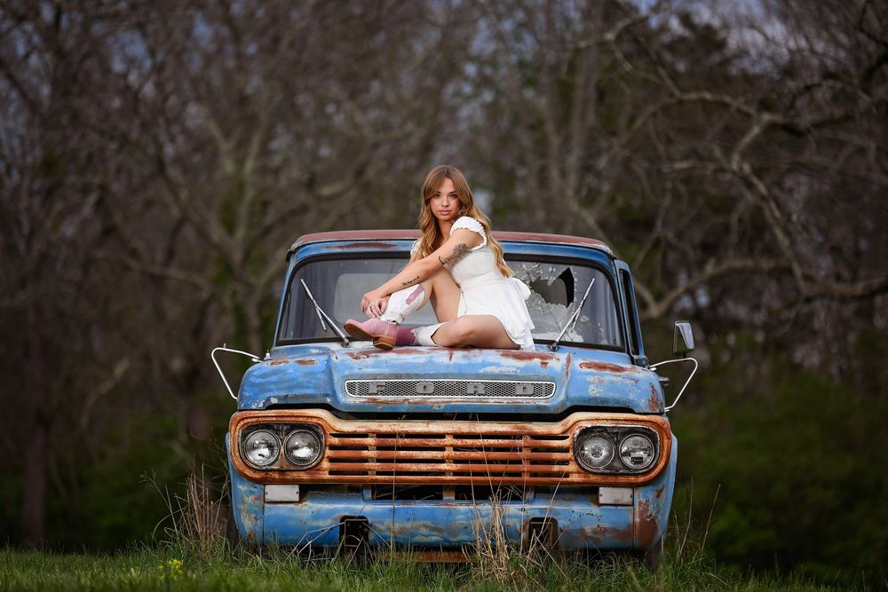 Young woman in white dress sitting on hood of rusty vintage blue Ford truck in a rural outdoor setting.