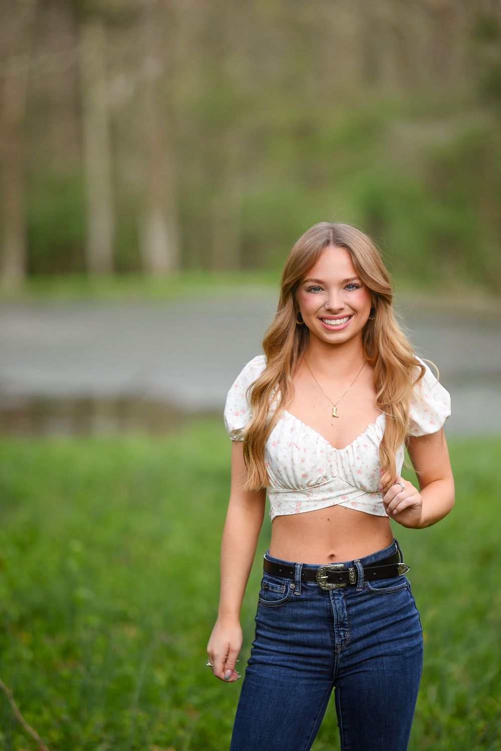 Young woman smiling outdoors in white crop top and jeans, standing on green grass with blurred trees background.