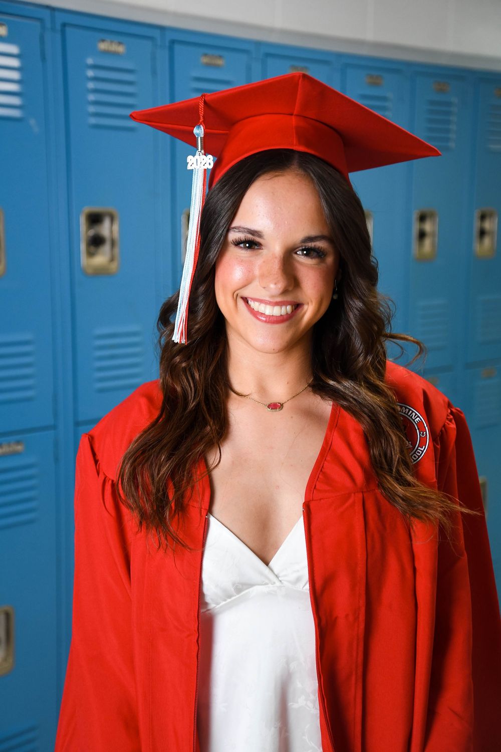 Smiling young woman in red graduation cap and gown posing in front of blue school lockers.