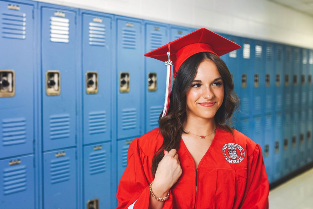 Smiling female graduate in red cap and gown posing in front of blue school lockers.