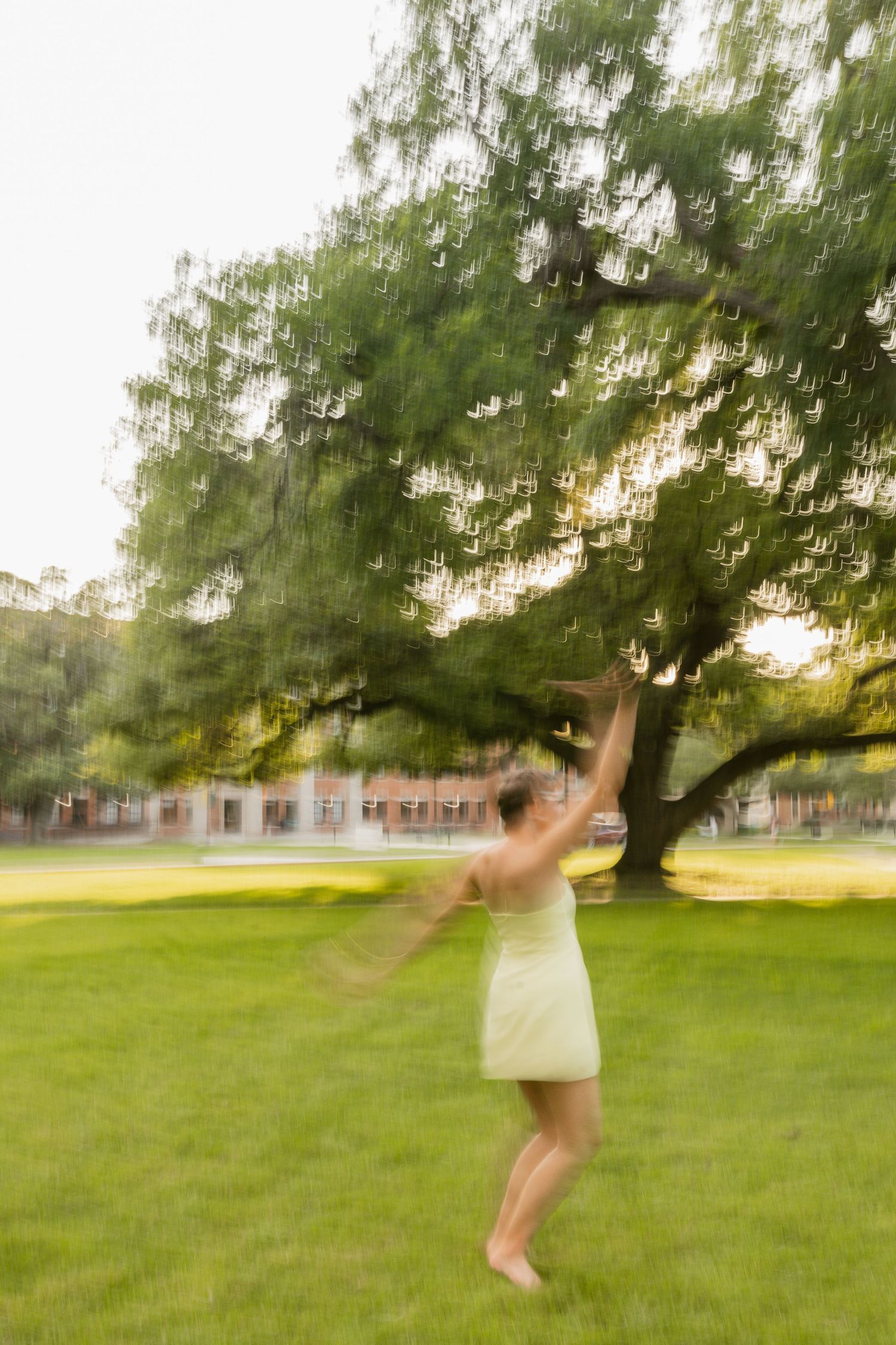 A woman in a white dress dances joyfully under a large green tree on a sunny day at Baylor University.