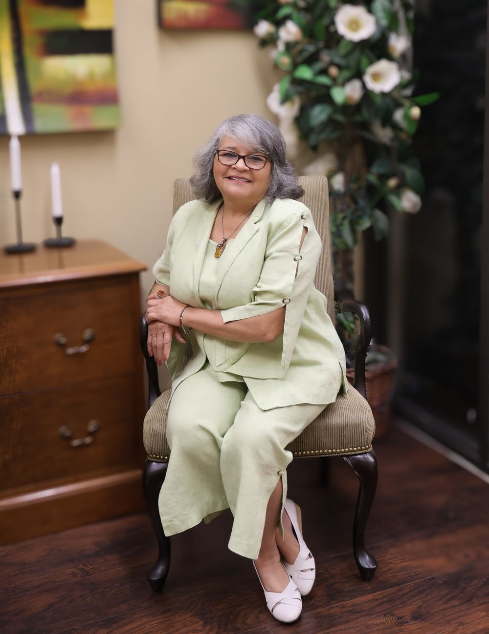A person in a light green suit sits elegantly in a chair with floral decor in the background.