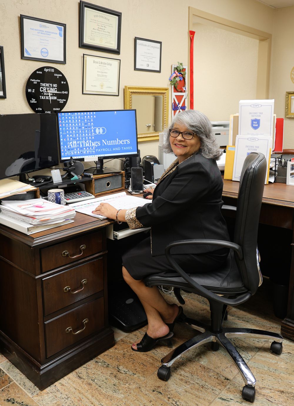 A professional seated at an office desk with computer monitors and certificates on the wall.