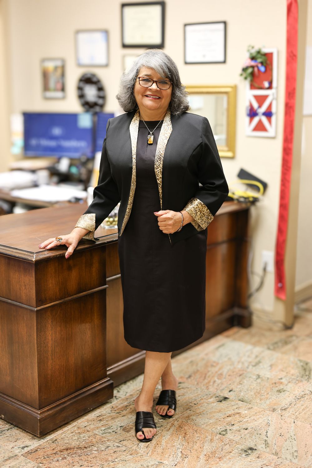 A professional in a black dress with gold accents poses in an office setting.