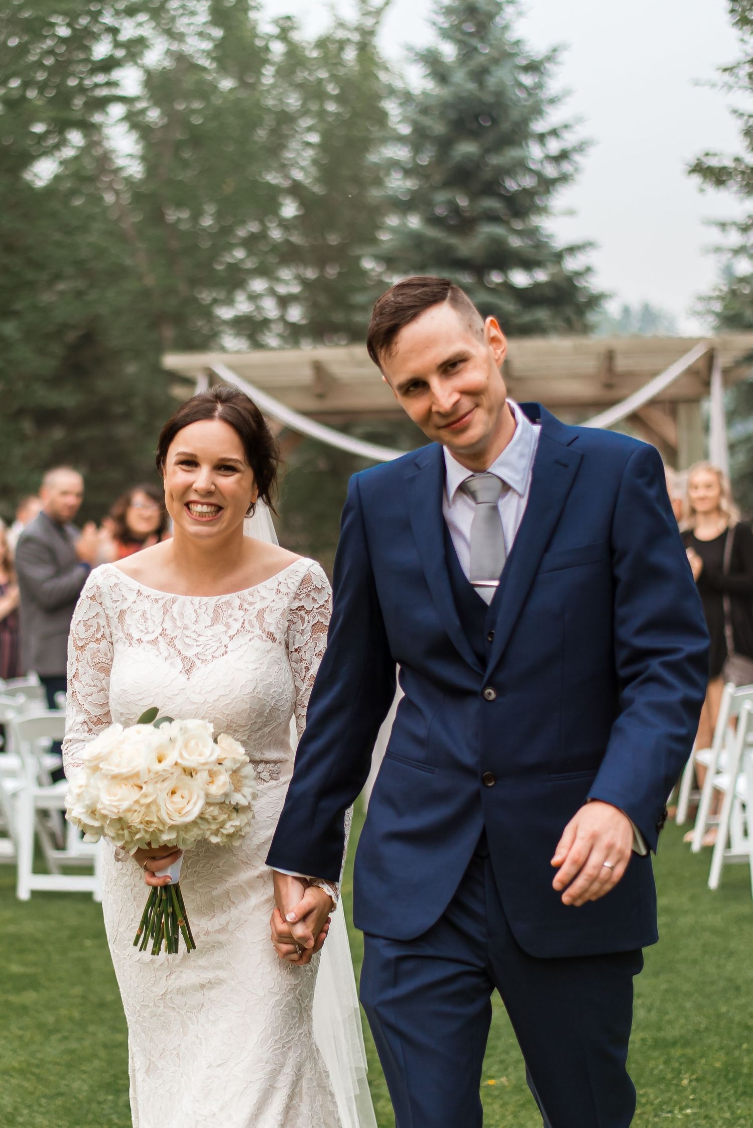 A newlywed couple walks hand in hand on grass with white chairs in the background.