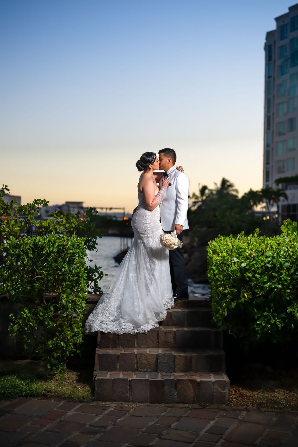 Elegant couple embraces on stone steps during sunset with lush greenery and high-rise building in background.
