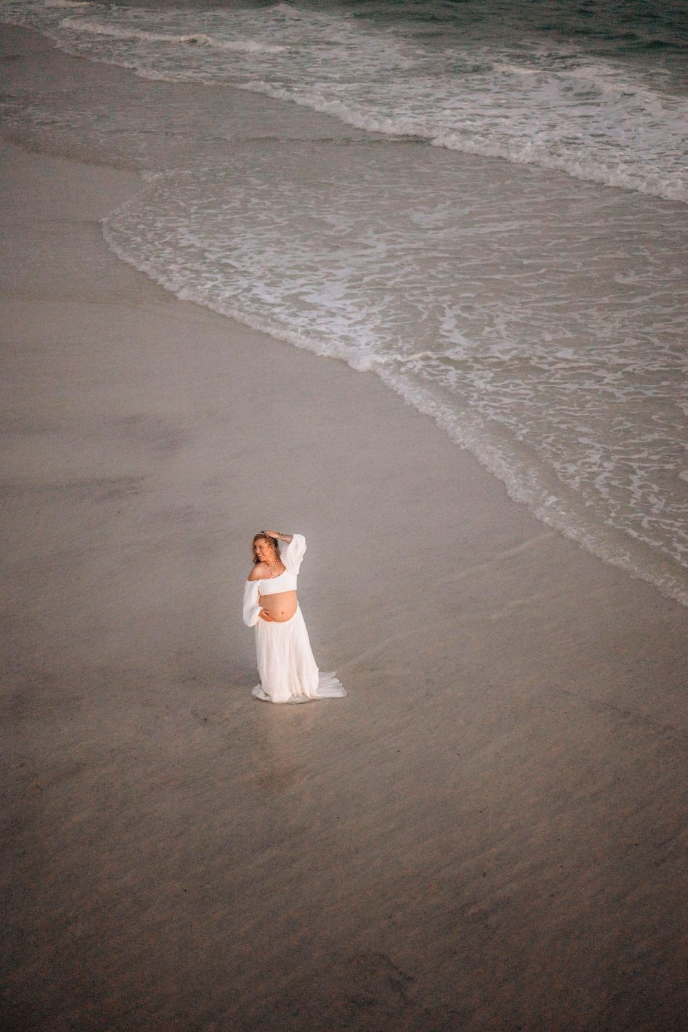 An aerial beach photograph captures a lone figure in white seated on the sandy shore.