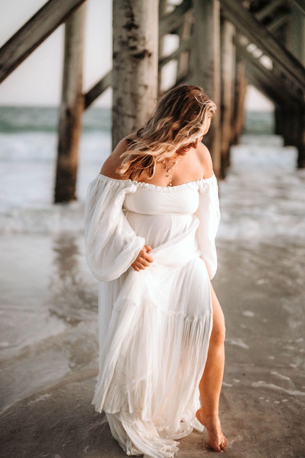 A flowing white dress catches the breeze beneath a pier as waves roll onto the beach.