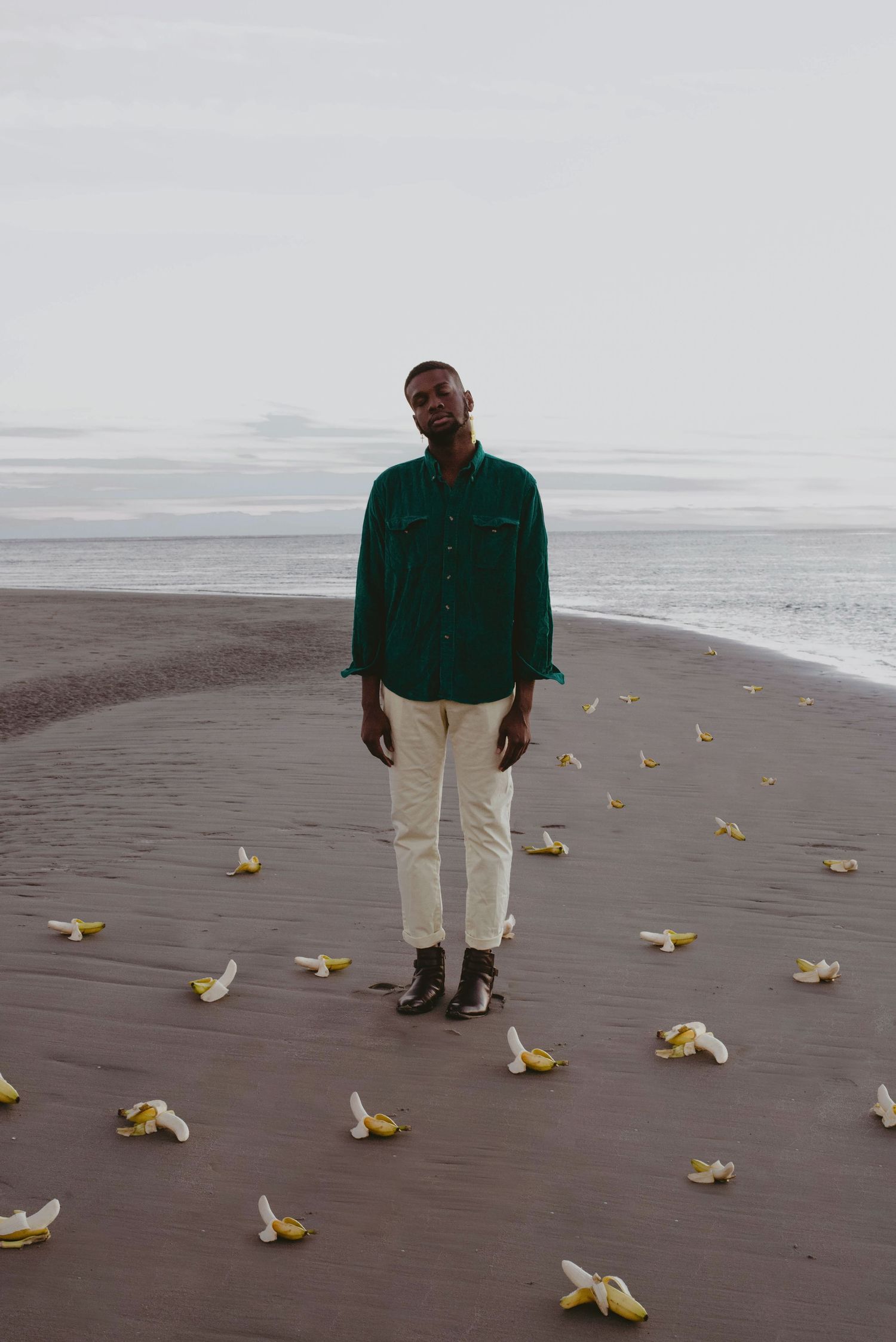 A portrait of a man standing on the beach with his eyes closed with bananas surrounding him on the ground.