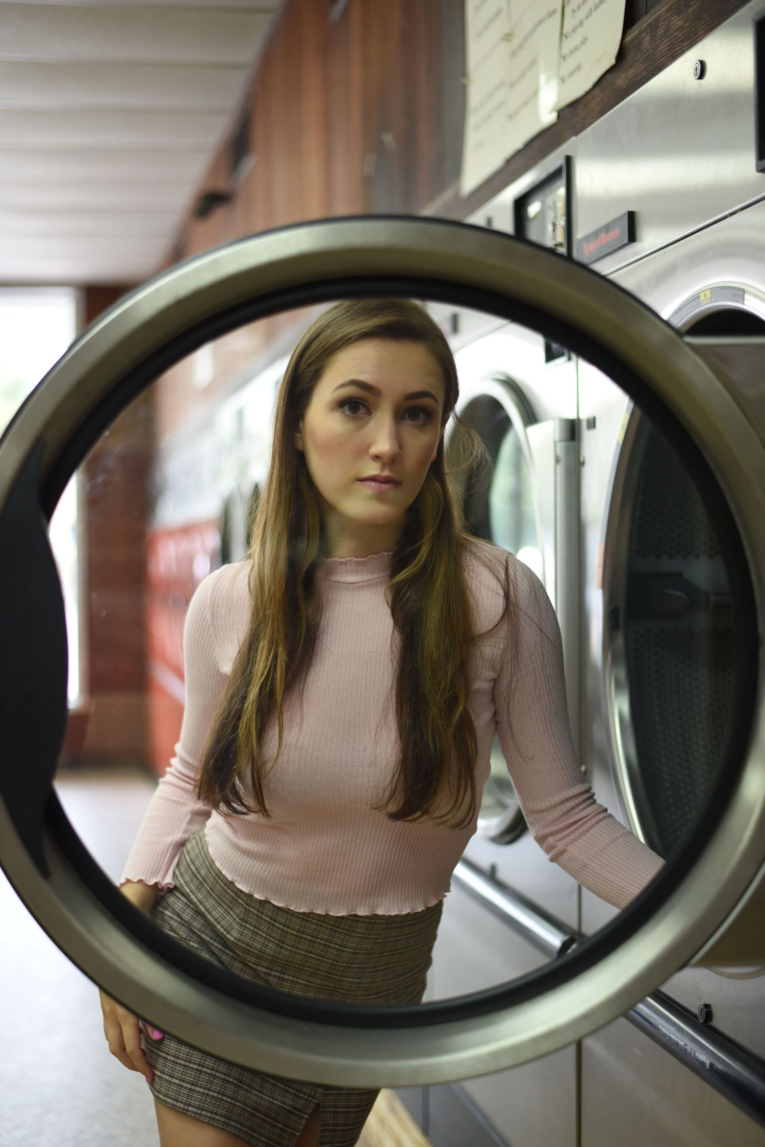 A portrait of a girl at a laundry mat posing behind the door of a washing machine.