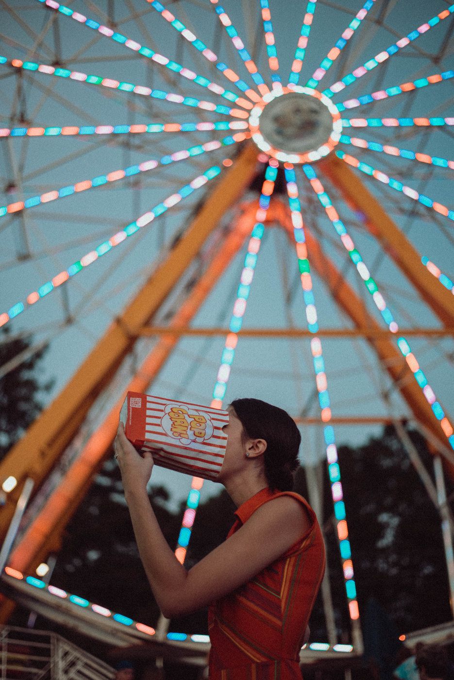 A portrait of a girl sticking her head in a bag of popcorn with the ferris wheel lit up behind her.