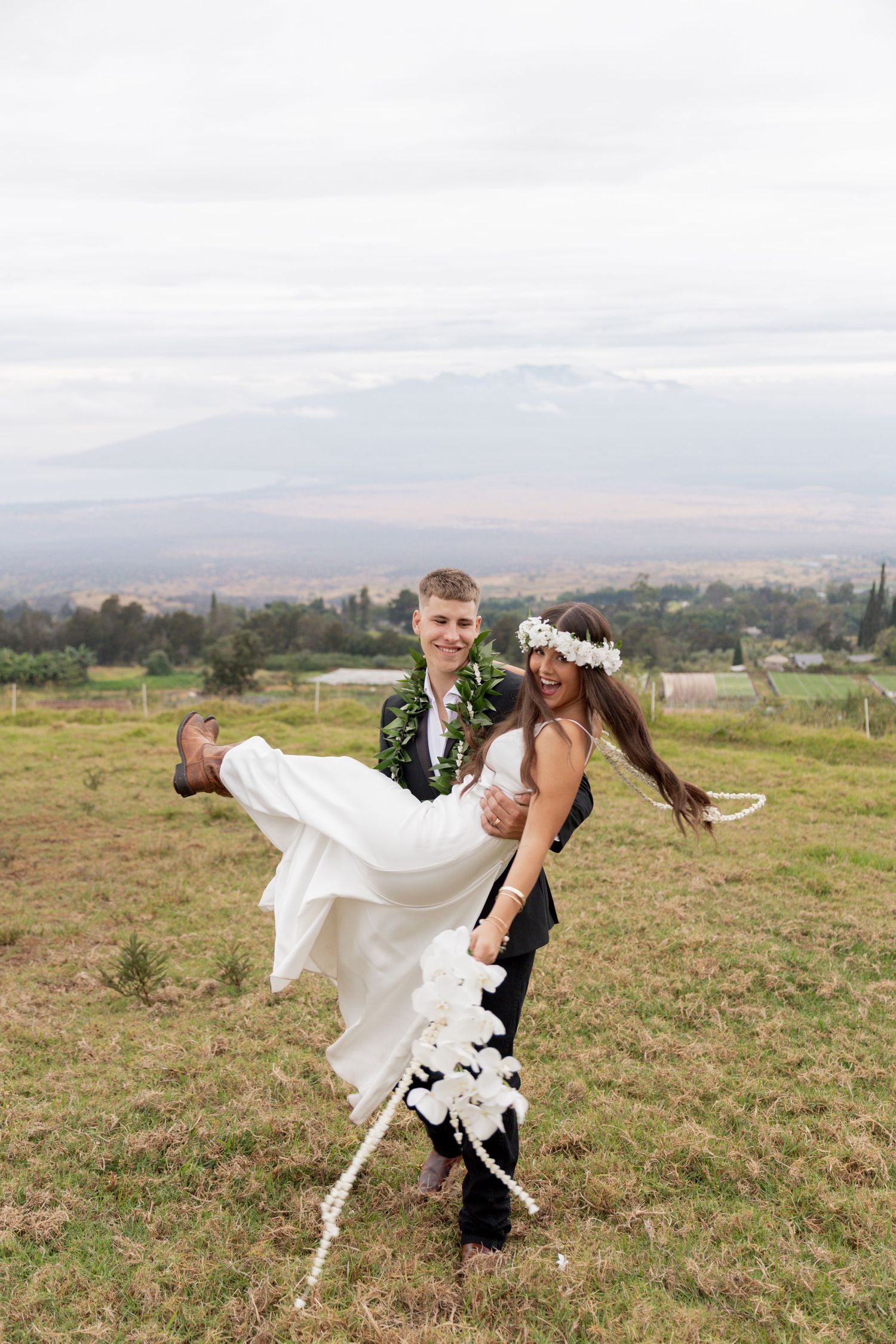 A couple in wedding attire dances playfully in a scenic mountain meadow with sweeping valley views in the background.