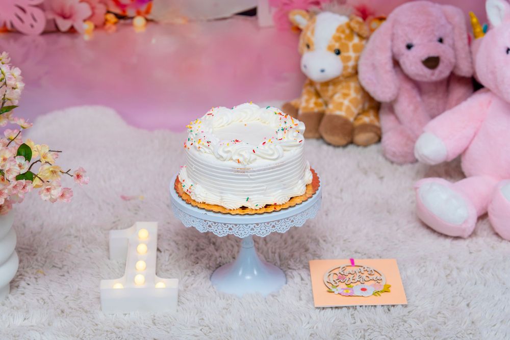 White birthday cake on blue stand with number one candle and pink plush toys in background.