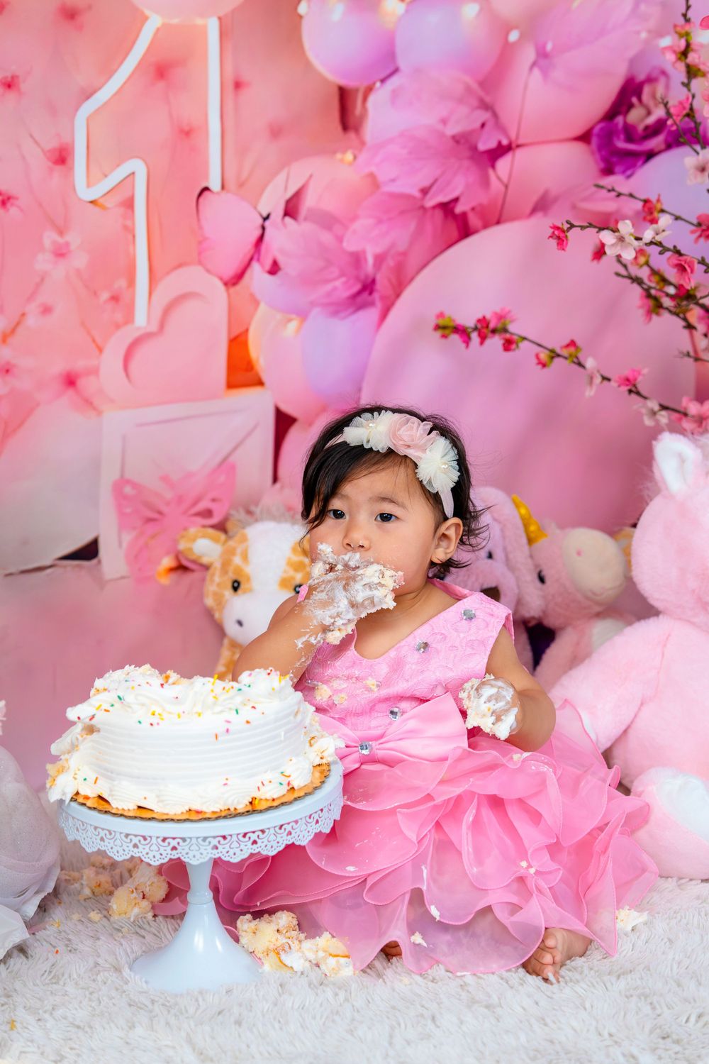 A first birthday cake smash photo with pink decorations and balloons in the background.