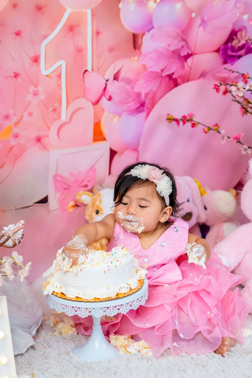 A baby in a pink dress enjoying their first birthday cake smash photoshoot.