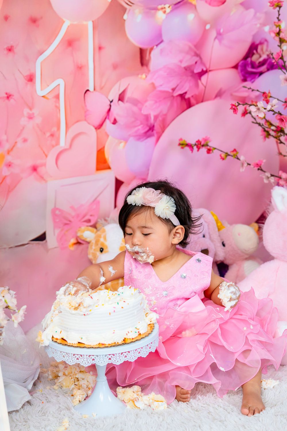 Baby girl in tulle dress having fun during cake smash session with pink balloon backdrop.