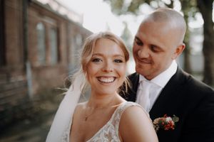 Smiling bride in lace wedding dress with groom nuzzling her cheek outdoors.