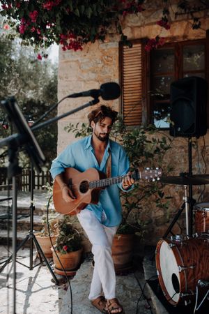 Musician playing acoustic guitar at an outdoor wedding reception near a stone building.
