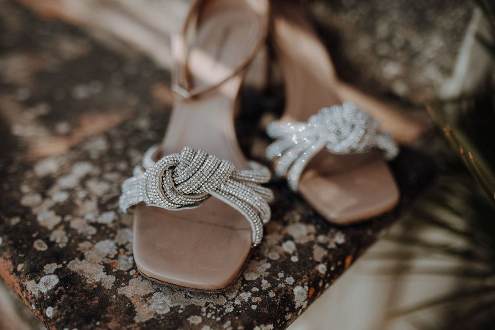 Elegant crystal knot-embellished bridal sandals displayed on rustic stone surface with palm leaf.