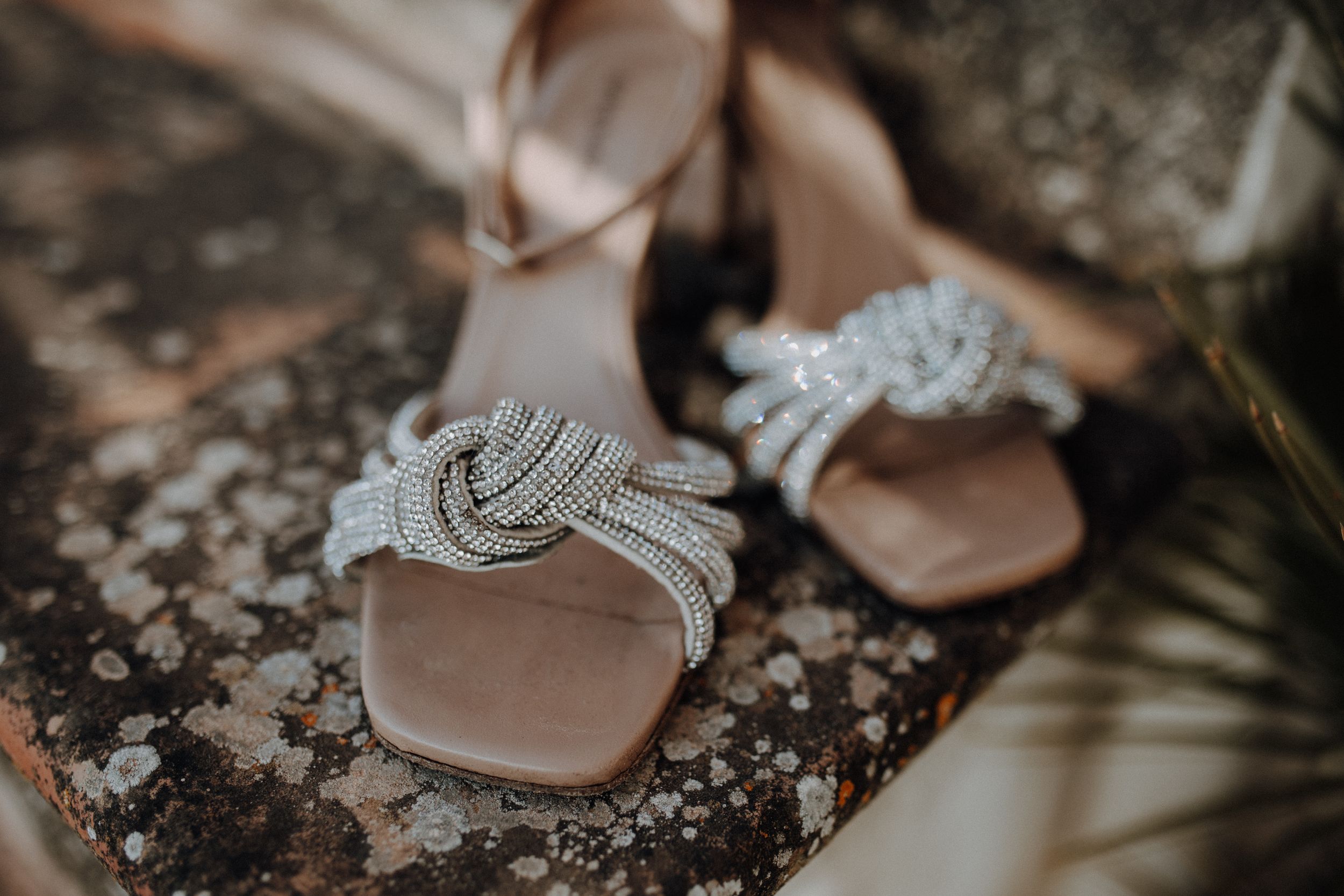 Elegant crystal knot-embellished bridal sandals displayed on rustic stone surface with palm leaf.