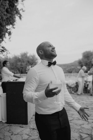 Bald groom in black tuxedo and bow tie celebrating joyfully at outdoor wedding reception.