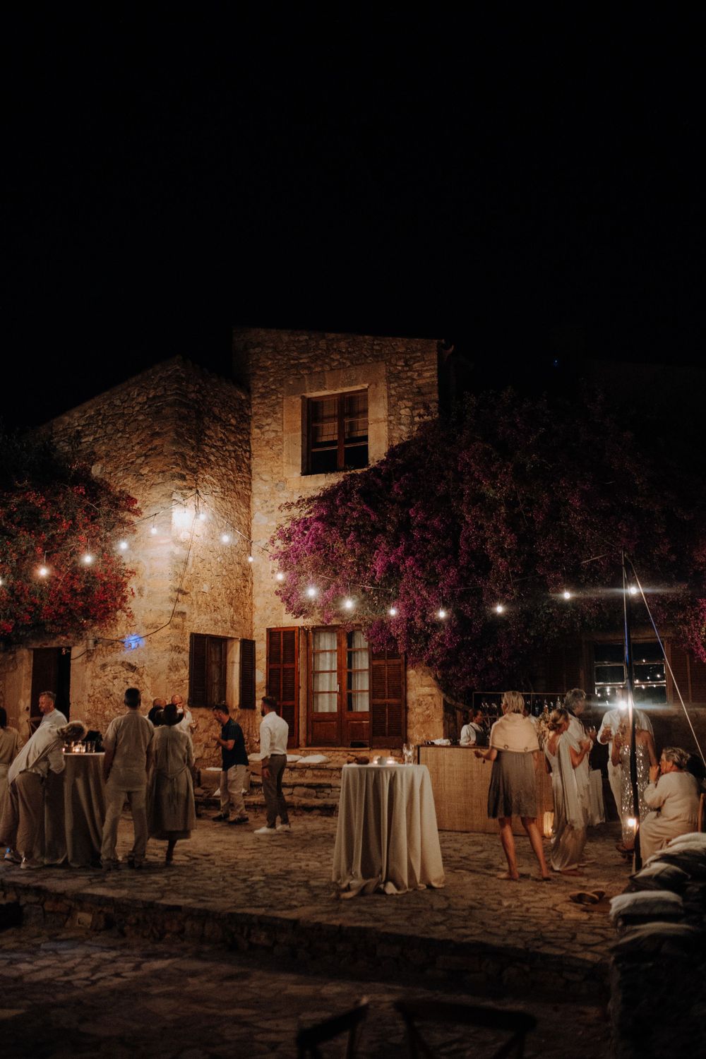 Guests mingling outside a rustic stone venue decorated with fairy lights at night.