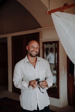 Smiling bald groom in white linen shirt gets dressed in warmly lit rustic room before wedding.