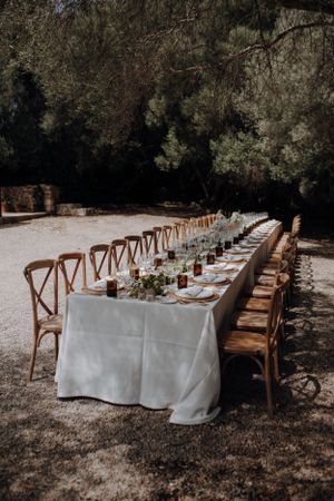 Long rustic outdoor wedding reception table set with white linen and greenery centerpieces.