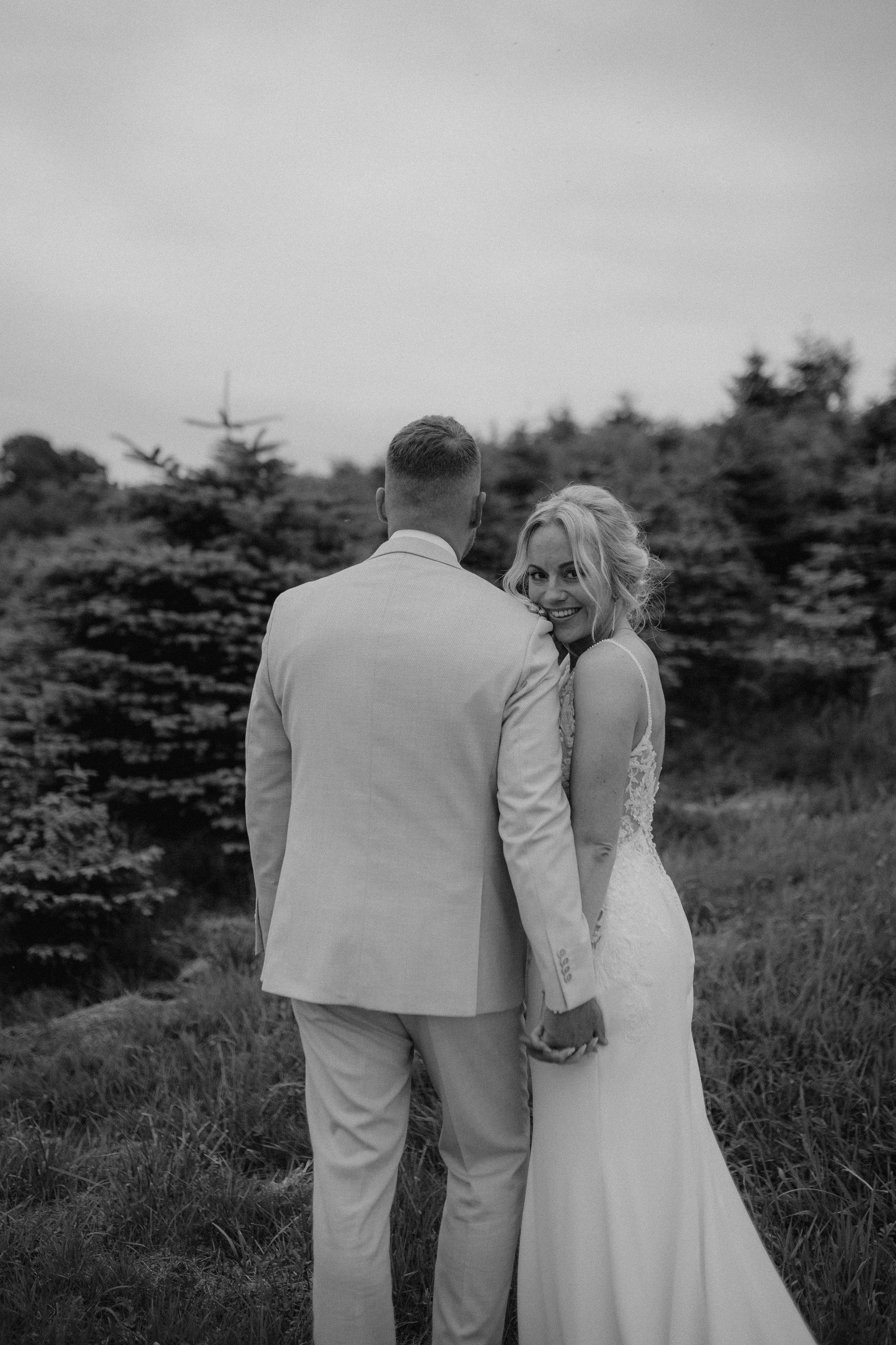 Black and white photo of bride looking back at camera while holding hands with groom outdoors.