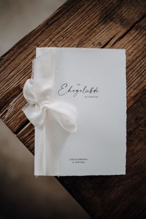 Elegant white wedding vow booklet tied with silk ribbon on rustic wood.