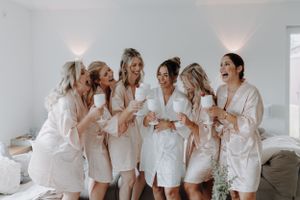 Bridesmaids in satin robes laughing and toasting with mugs before wedding.