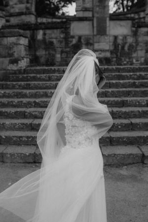 Black and white photo of bride in lace gown and flowing veil near stone steps.