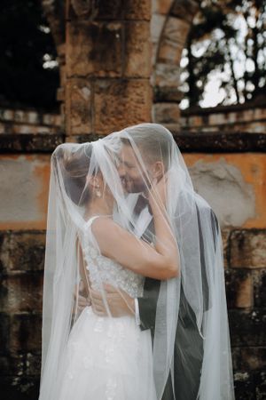 Bride and groom sharing intimate moment under flowing veil at stone ruins.